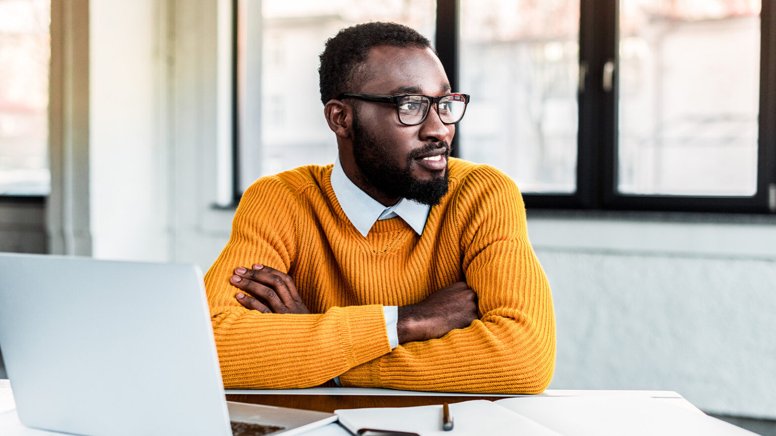Man with glasses and beard in a yellow sweater sits at a desk with a laptop, looking to the side in a bright office space.