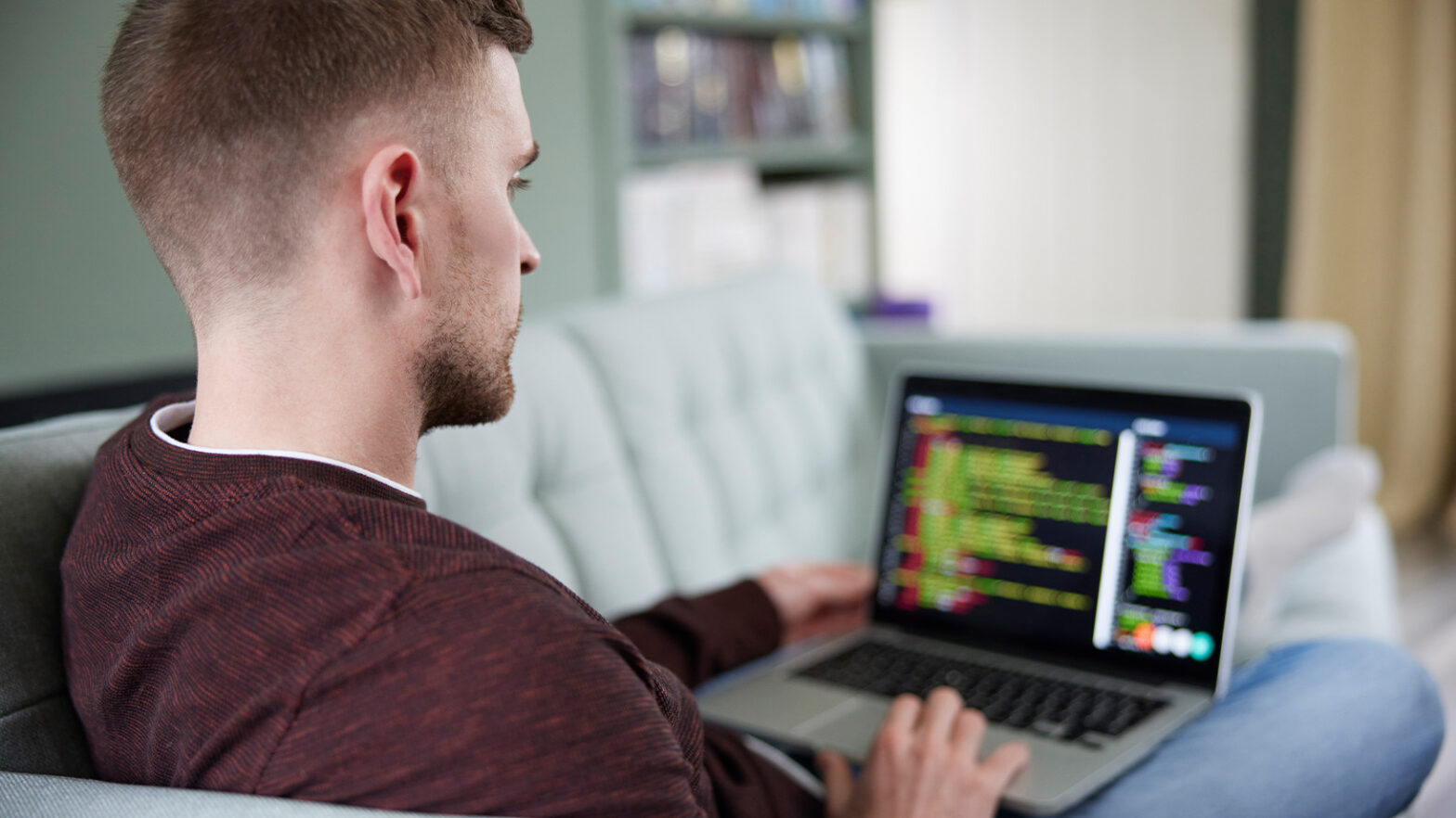 A person sitting on a sofa is using a laptop with code on the screen, focused on the work, in a room with a bookcase.