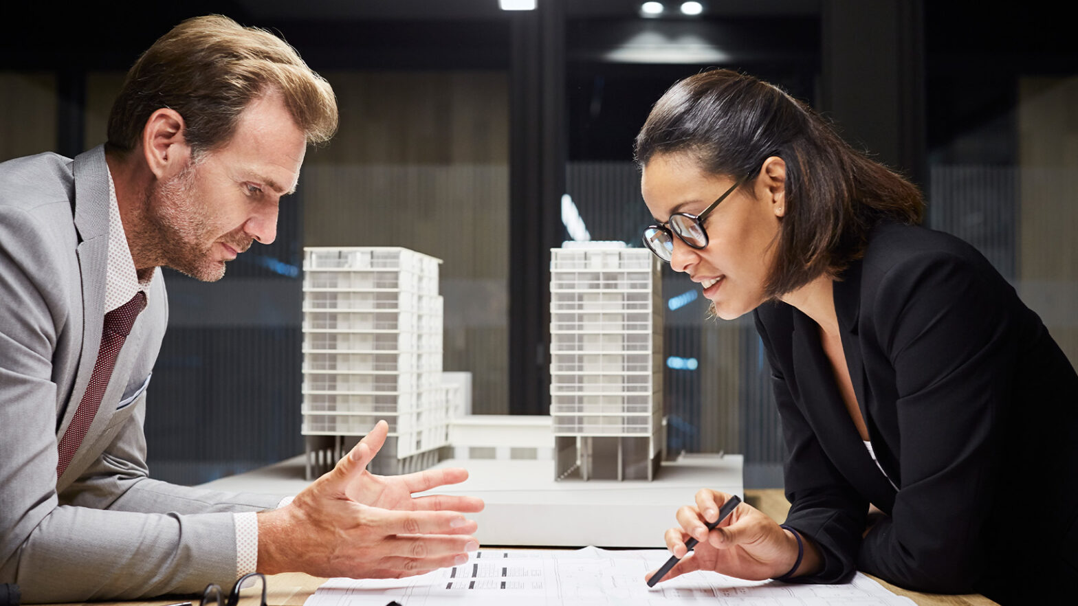 A man and woman in business attire discuss architectural plans at a table with building models in the background.