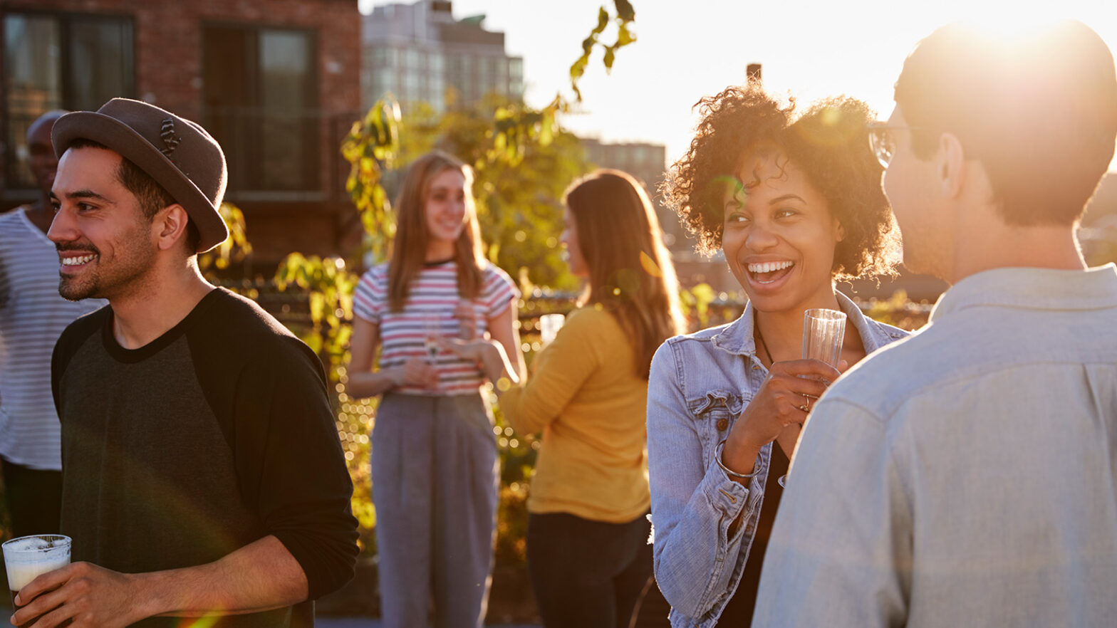 People at a rooftop gathering, smiling and holding drinks. The sun sets in the background, casting a warm glow over the scene.