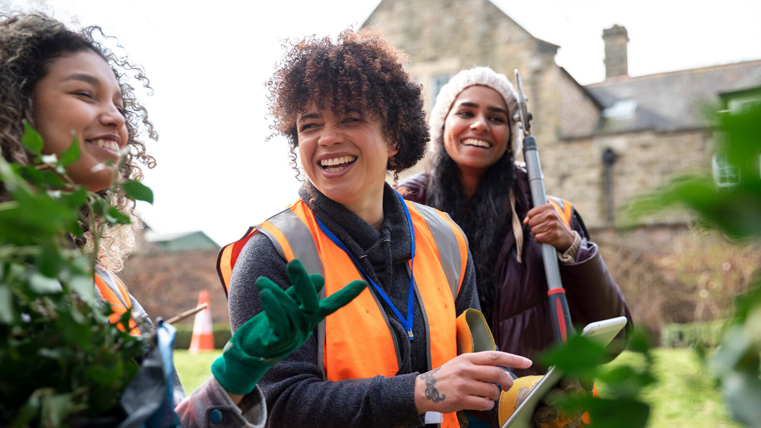 Three people wearing orange safety vests garden joyfully together, holding tools, with buildings in the background.