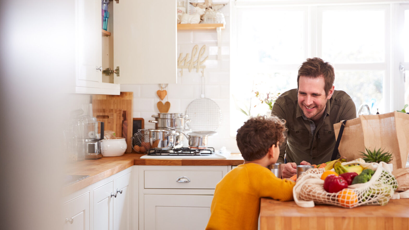 A man and a child smile at each other in a bright kitchen. A paper bag and various fruits and vegetables are on the counter.