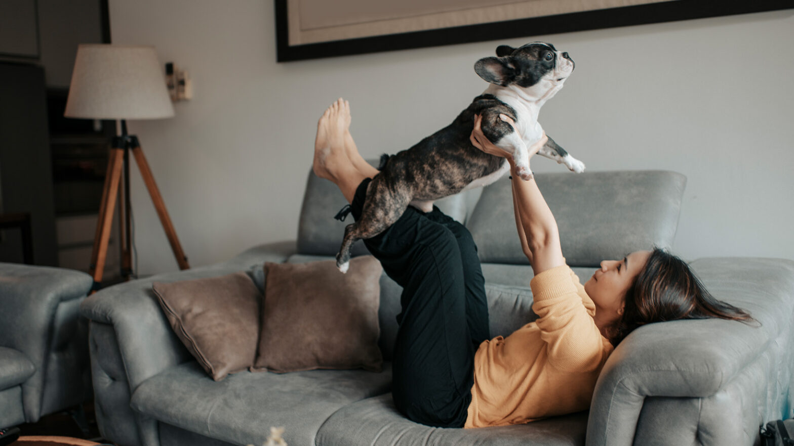 Person lying on a sofa, lifting a black and white dog with their legs and arms. The room has a lamp and cushions.