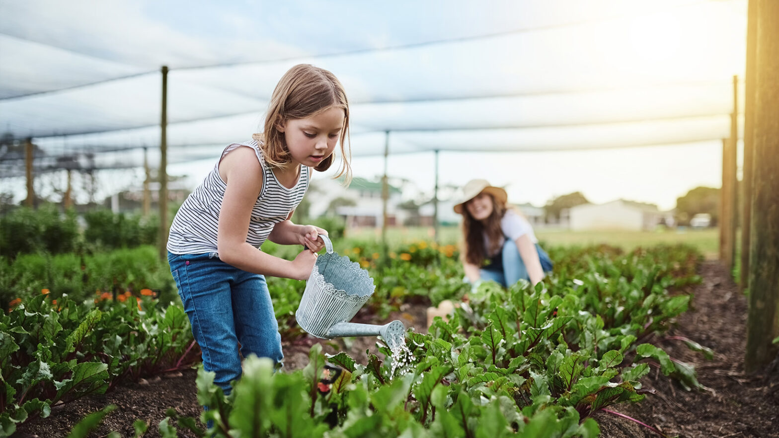 A young girl waters plants with a watering can in a garden, while another person tends to plants in the background.