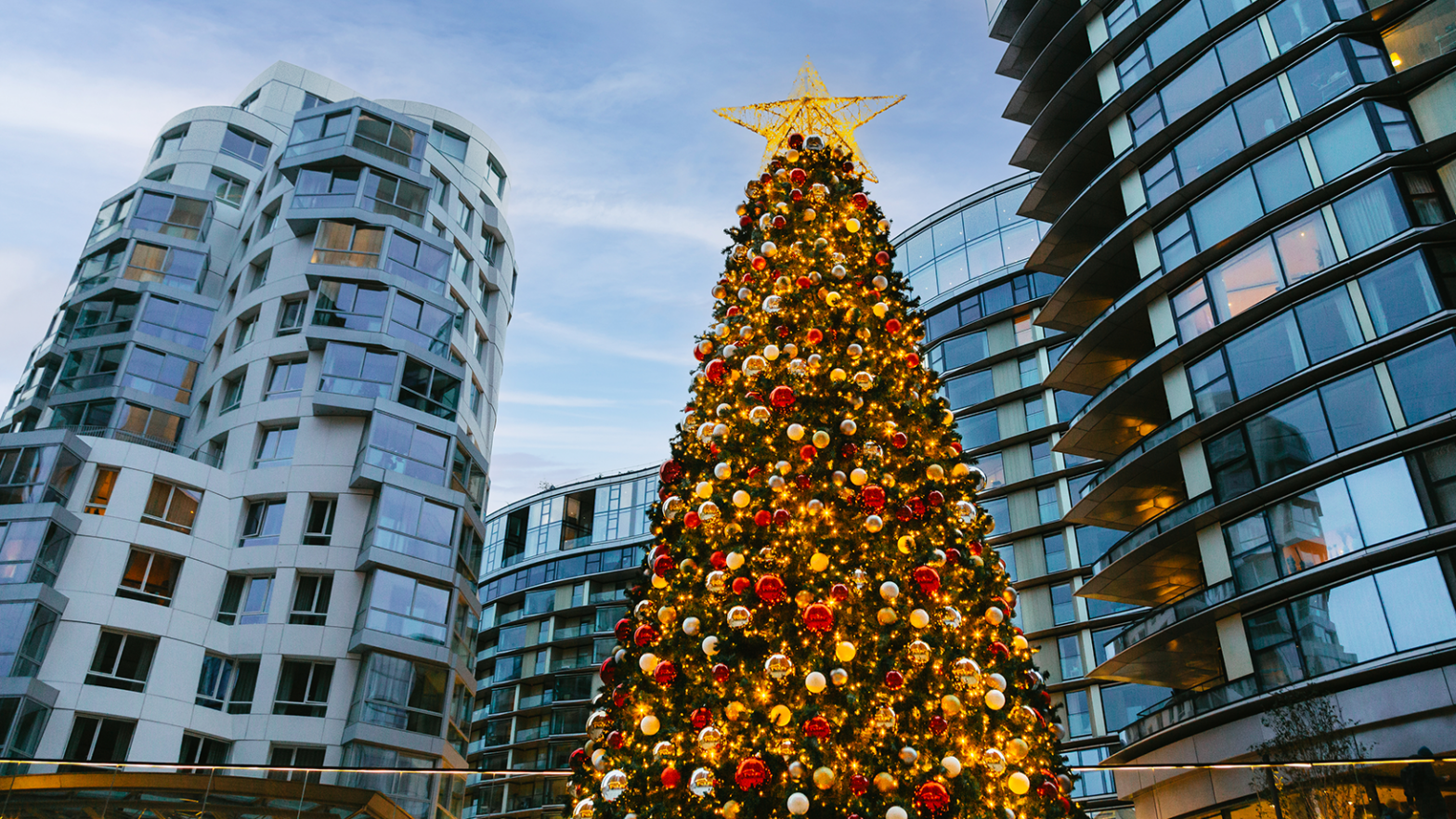 A large decorated Christmas tree with a star on top is surrounded by modern, curved glass buildings under a cloudy sky.