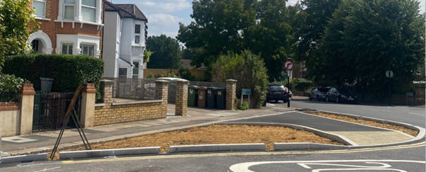 A newly constructed curved curb and sidewalk bordered by trees and houses, with some japanese knotweed nearby.